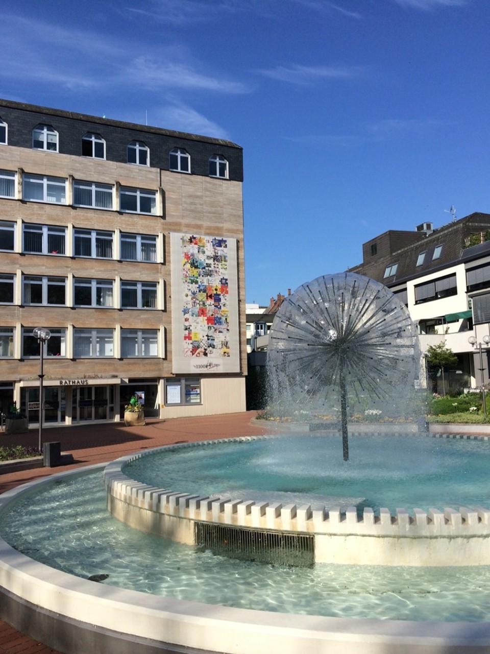Deutschland - Limburg an der Lahn - Der Brunnen auf dem Serenadenhof, dem Platz vor der Stadthalle von Limburg, wird von den Einwohnern bisweilen auch als „Pusteblume” bezeichnet. Als sogenannte „puffball fountain” besteht er aus einer Metall-Säule an deren Ende 127 Düsen eine Kugel bilden. Damit erinnert der Brunnen von seiner Form her an den verblühten Löwenzahn. „Stadthallenbrunnen” ist eine weitere Bezeichnung für das Wasserspiel auf dem Serenadenhof. Die meisten 
Stadtführungen durch Limburg beginnen bei der „Pusteblume”. Der Brunnen gilt als ein Wahrzeichen der Stadt und ist zudem das Firmenemblem der Stadthallen GmbH.