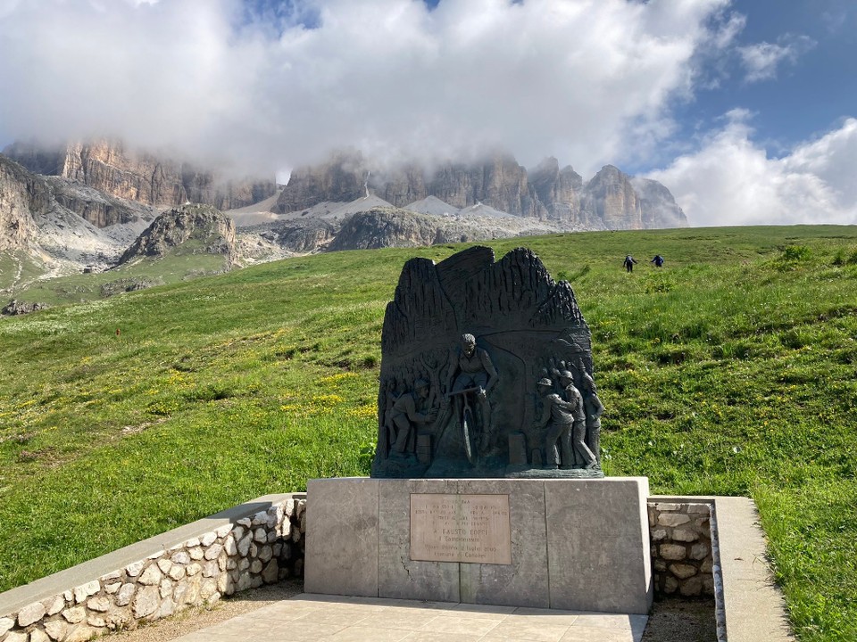 Italien - Livinallongo del Col di Lana - Die Sella zeigt sich hinter einem Denkmal für einen Radrennfahrer.